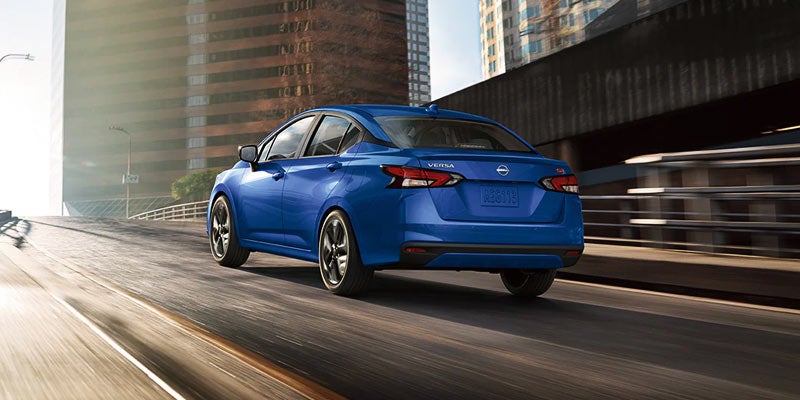 Rear angled view of a 2024 blue Nissan Versa driving on road past buildings
